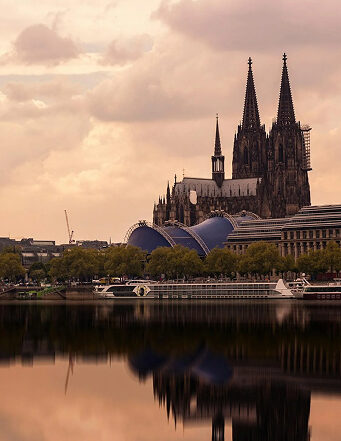 xuits-Kölner Dom mit zwei Türmen und Rhein im Vordergrund, Boote und blaue Struktur am Ufer.