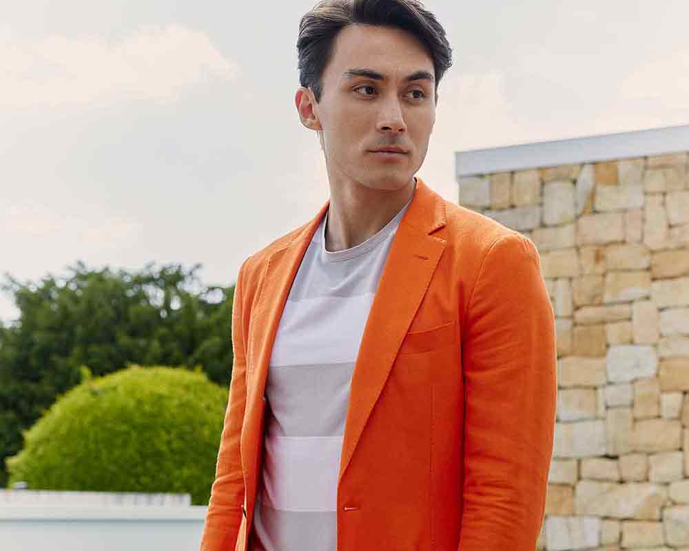 xuits -Mann mit orangefarbenem Blazer und weißem T-Shirt vor Steinmauer und grünen Büschen.