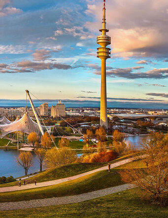 xuits-Olympiaturm und Olympiapark in München mit See und herbstlichen Bäumen bei Sonnenuntergang.