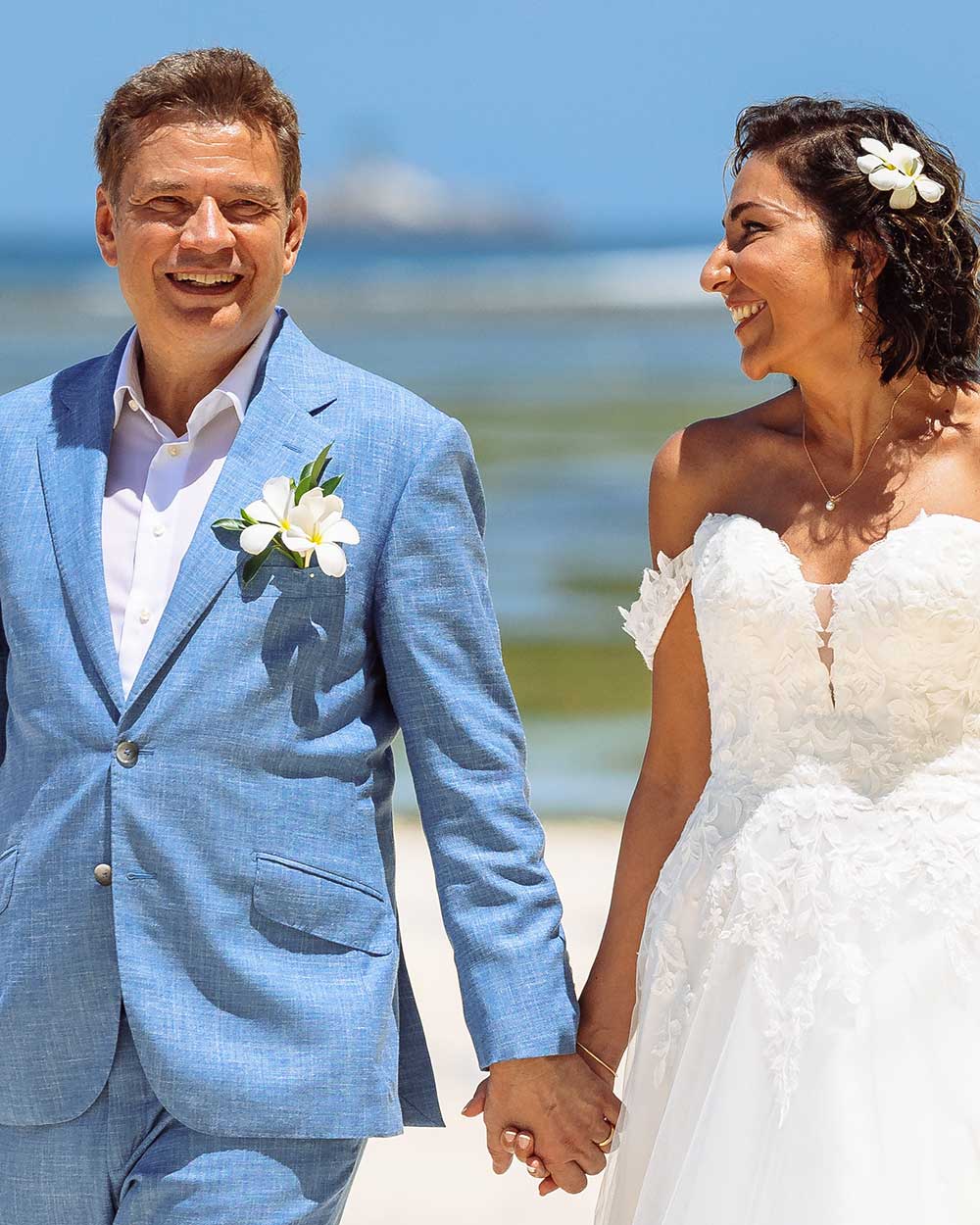 xuits-Brautpaar hält Händchen am Strand, Mann im hellblauen Anzug, Frau im weißen Spitzenkleid.