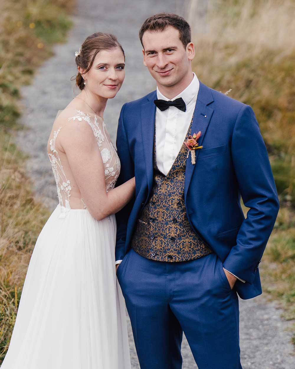 xuits-Brautpaar auf einem Weg im Freien, Braut in weißem Kleid, Bräutigam in blauem Anzug mit Weste.