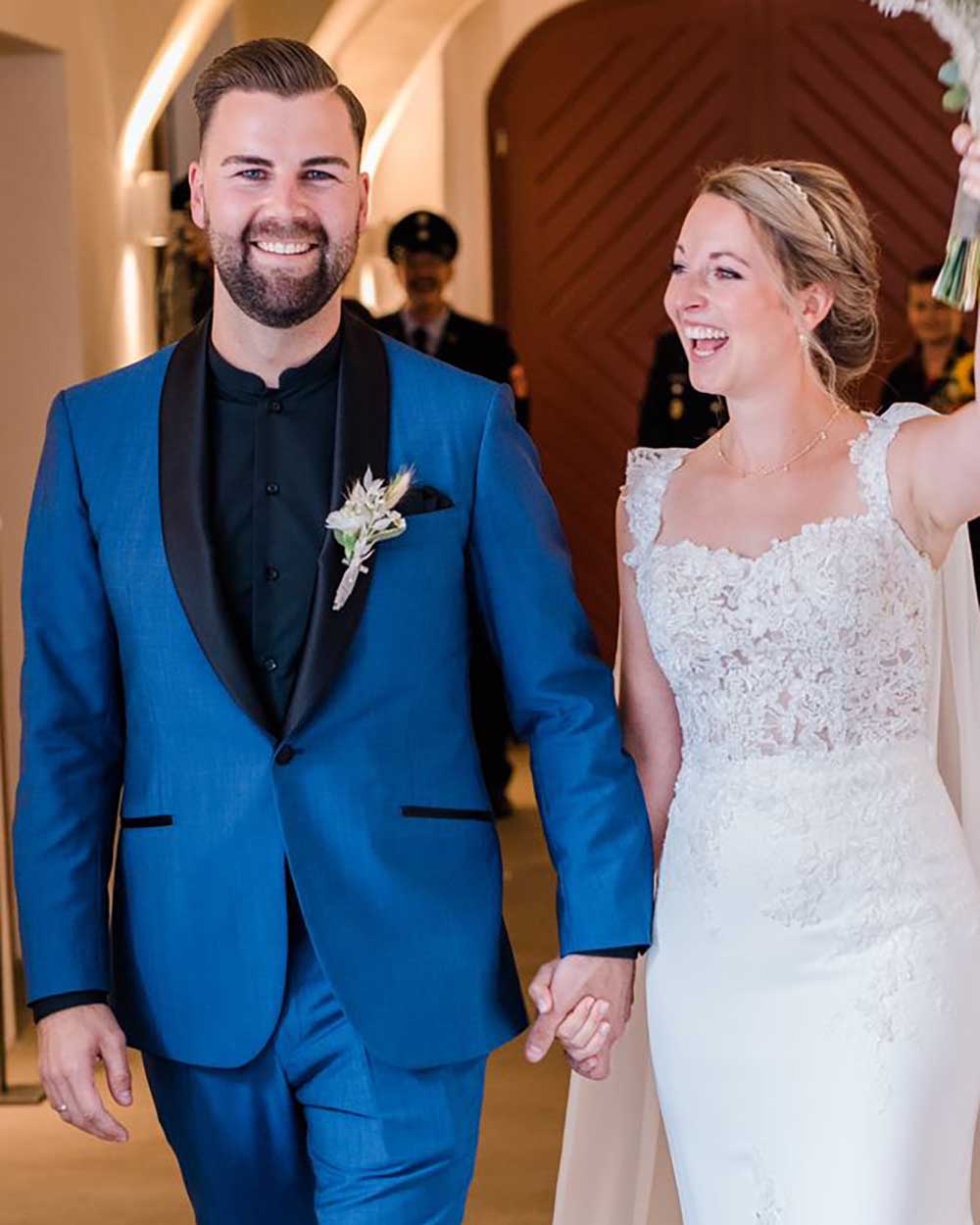 xuits-Brautpaar mit blauem Anzug und weißem Spitzenbrautkleid hält Händchen vor Holztür.