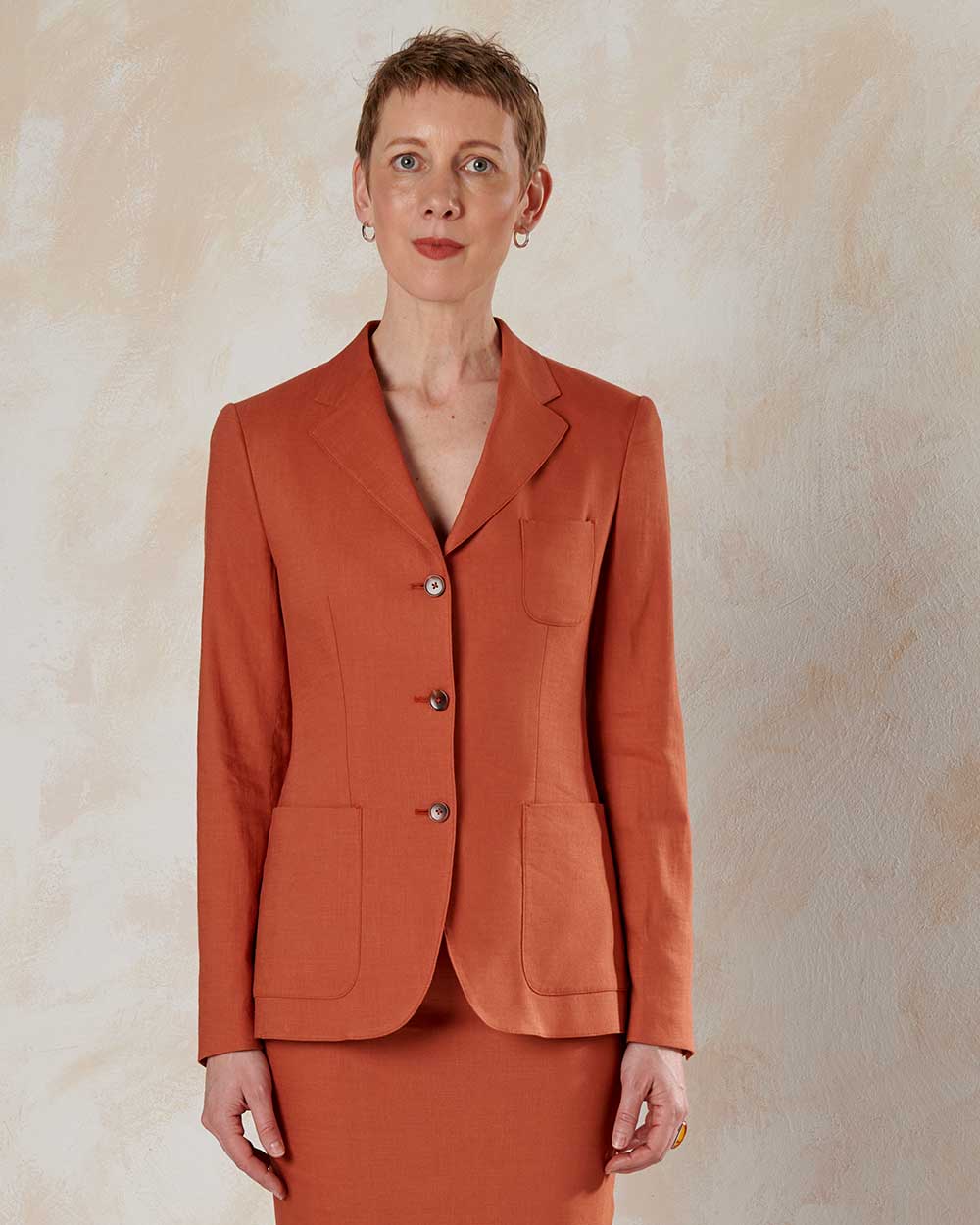 xuits-Frau mit kurzem Haar trägt orangefarbenen Blazer mit drei Knöpfen und aufgesetzten Taschen vor hellem Hintergrund.