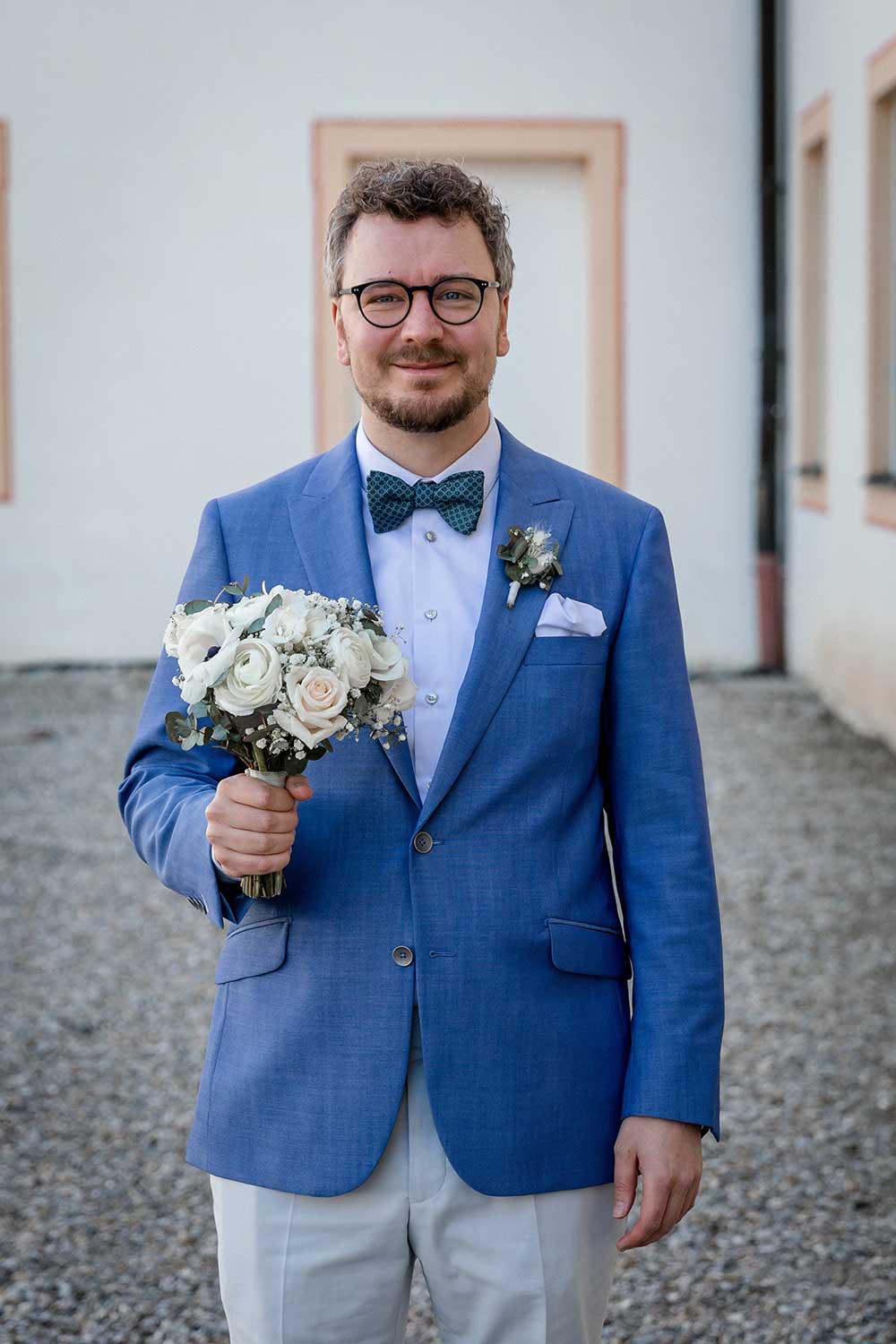 xuits-Mann im blauen Anzug mit Fliege hält Brautstrauß aus weißen und rosa Blumen vor hellem Gebäude.