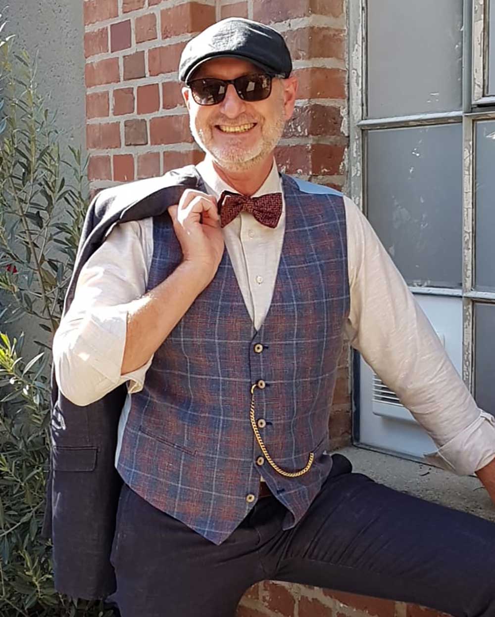 xuits -Mann in Hemd, Weste, Fliege, Flatcap und Sonnenbrille lehnt an Backsteinmauer mit Jackett über der Schulter.