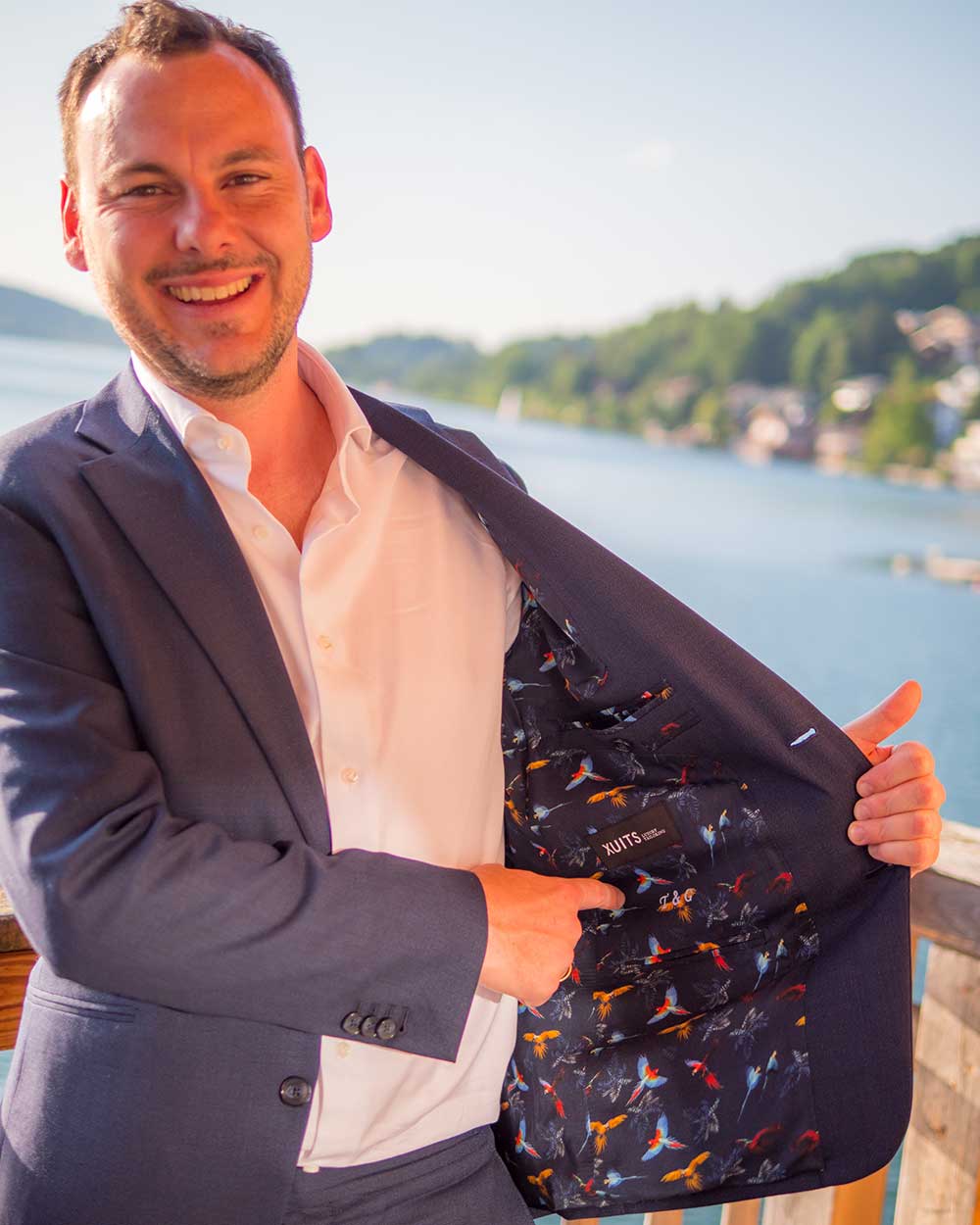 xuits-Mann im Anzug zeigt das bunte Papageienmuster des Futterstoffs seines Jacketts am Seeufer.