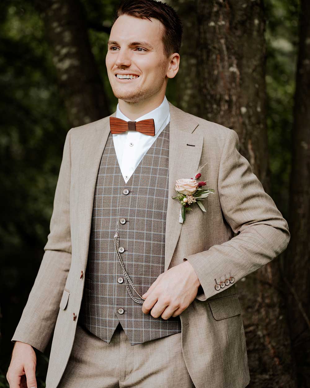xuits-Mann in beige Anzug mit karierter Weste, Holzbinder, Taschenuhr und Blumenanstecker vor Baum.