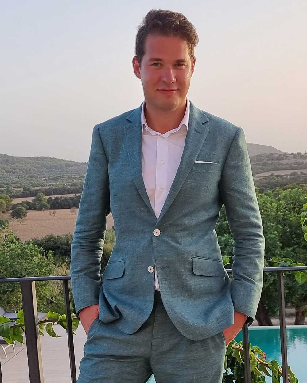 xuits-Mann im blauen Anzug steht vor Landschaft mit Bäumen, Feldern und Pool.