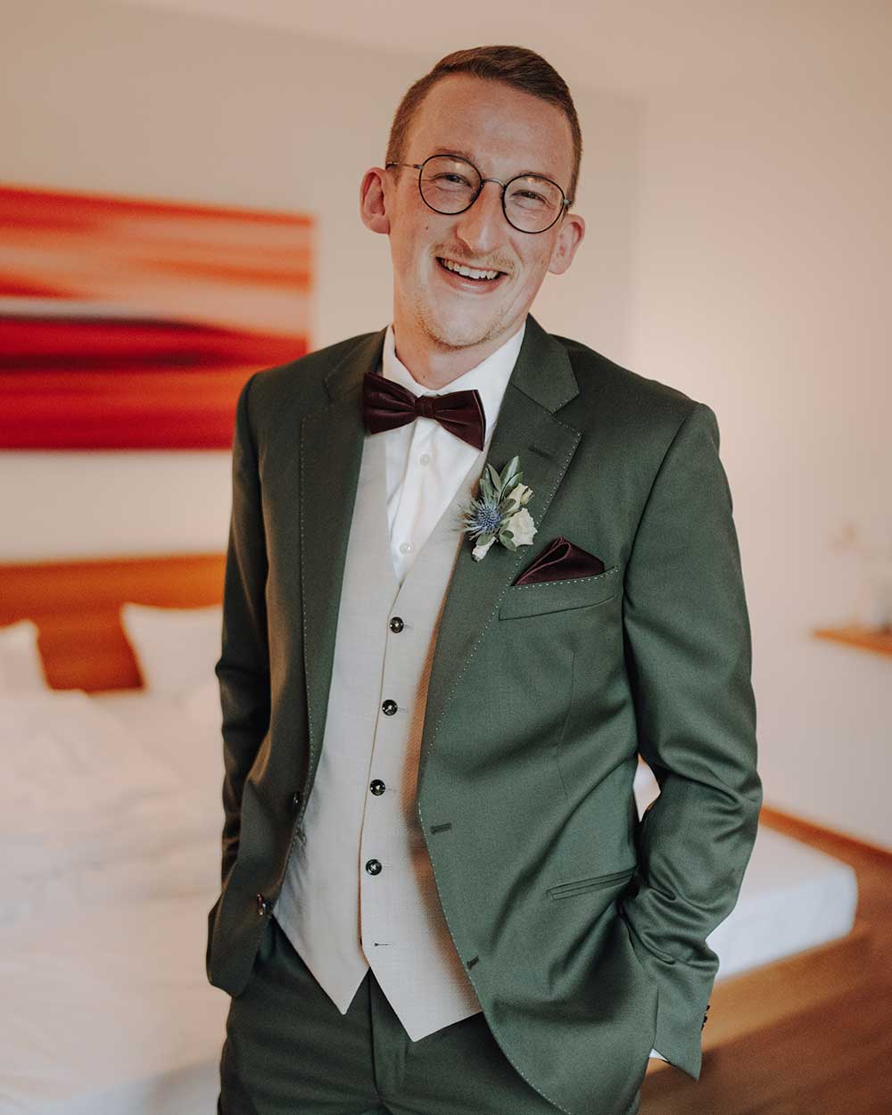 xuits-Mann mit Brille im dunkelgrünen Anzug, Fliege und Blumenanstecker in einem Zimmer mit Bett und Kunstwerk.