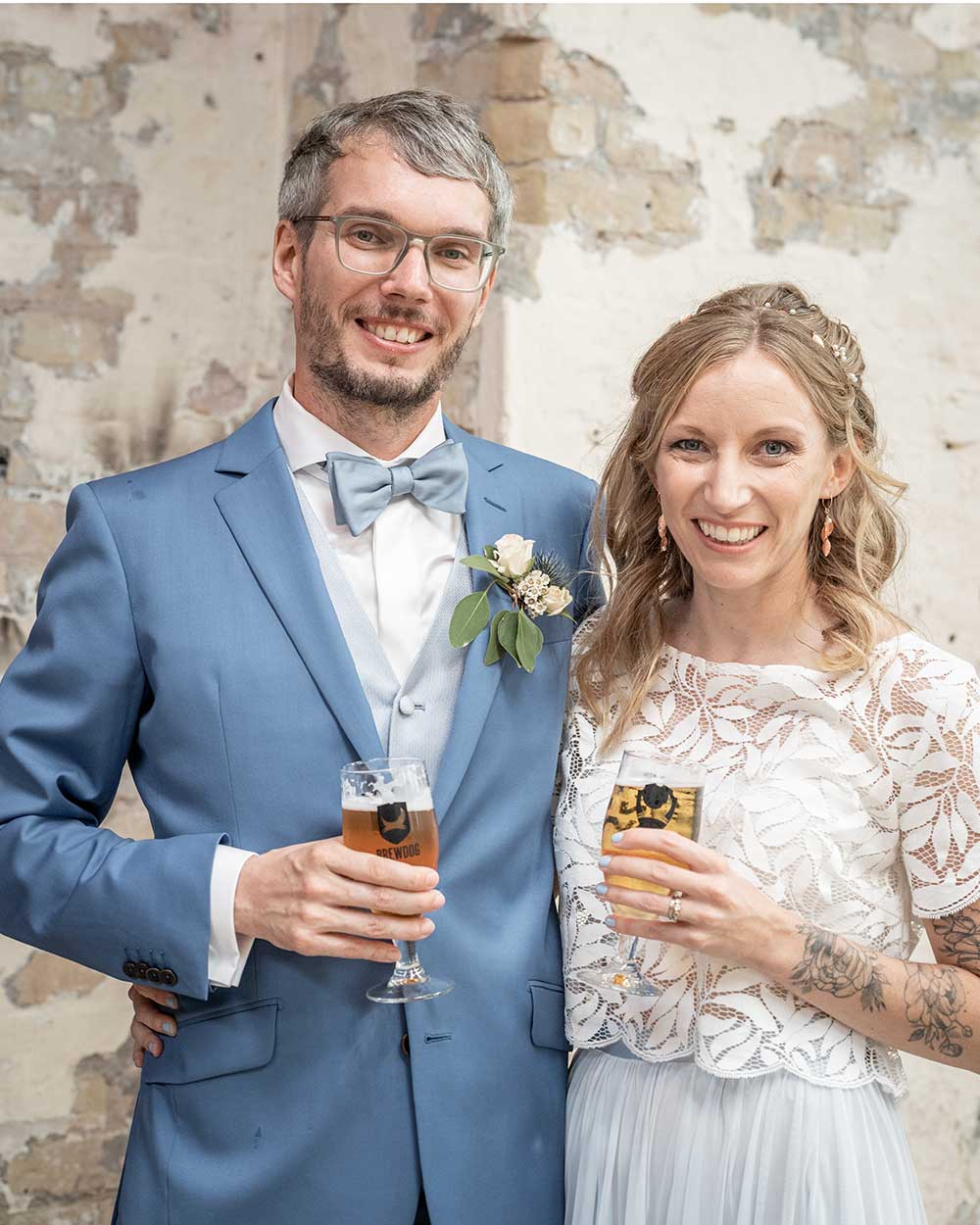 xuits-Brautpaar lächelnd mit Biergläsern vor verwitterter Mauer in Hochzeitskleidung.