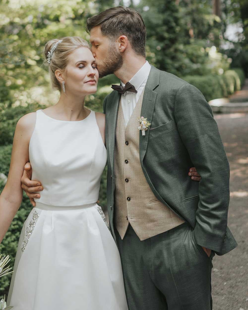 xuits-Brautpaar im Freien, Bräutigam küsst Braut auf die Stirn, Brautkleid weiß, Anzug mit Weste und Fliege.