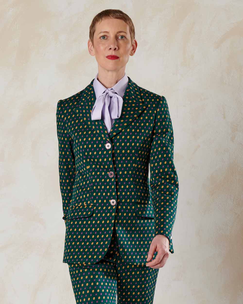 xuits-Person mit kurzem blondem Haar in grünem Anzug mit kleinem Blumenmuster und hellvioletter Bluse mit Schleife.