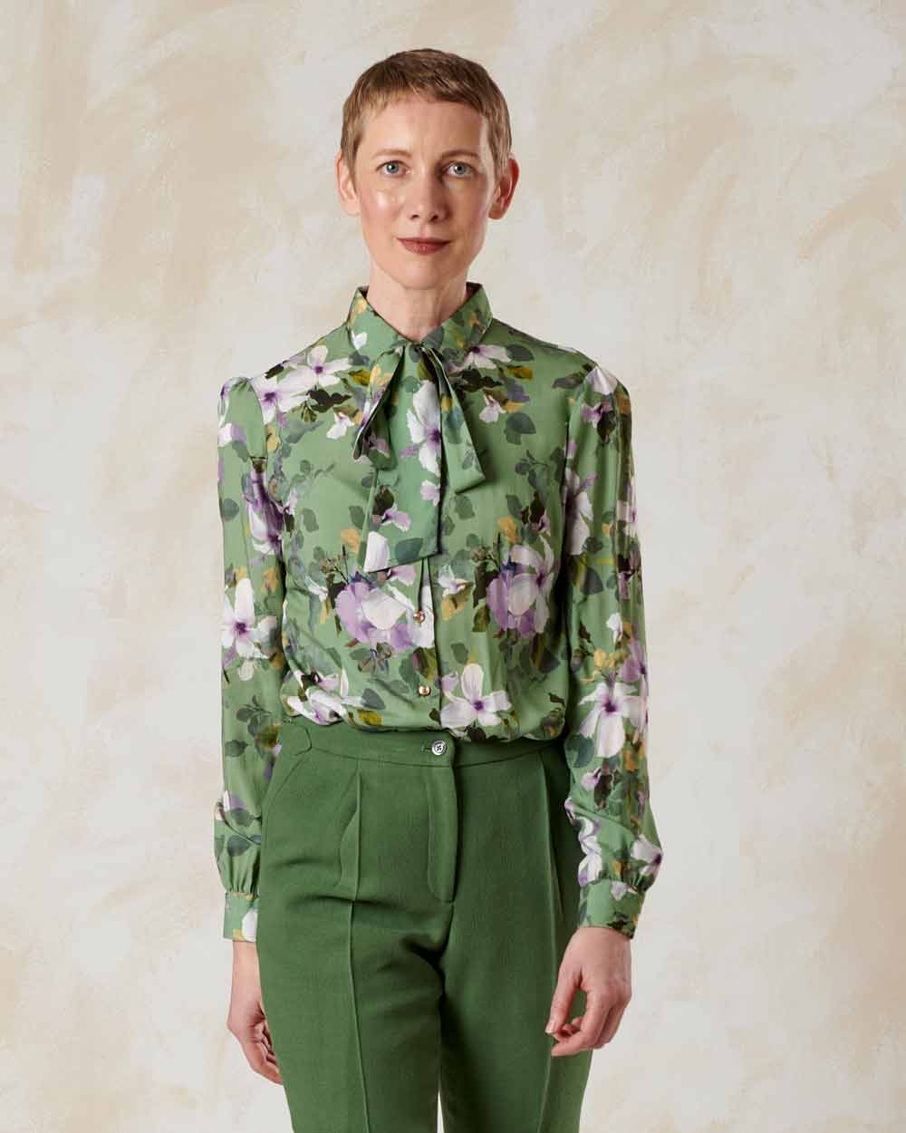 xuits -Frau mit kurzen Haaren in grüner, floral gemusterter Bluse mit Fliege und grüner Hose vor hellem Hintergrund.