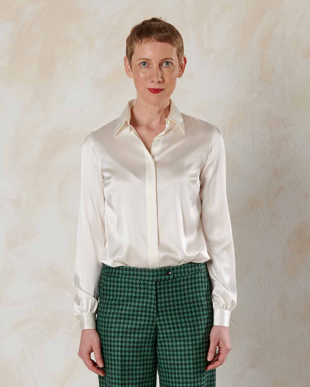 xuits -Frau mit kurzen Haaren trägt glänzende cremefarbene Bluse und grüne gemusterte Hose vor hellem Hintergrund.