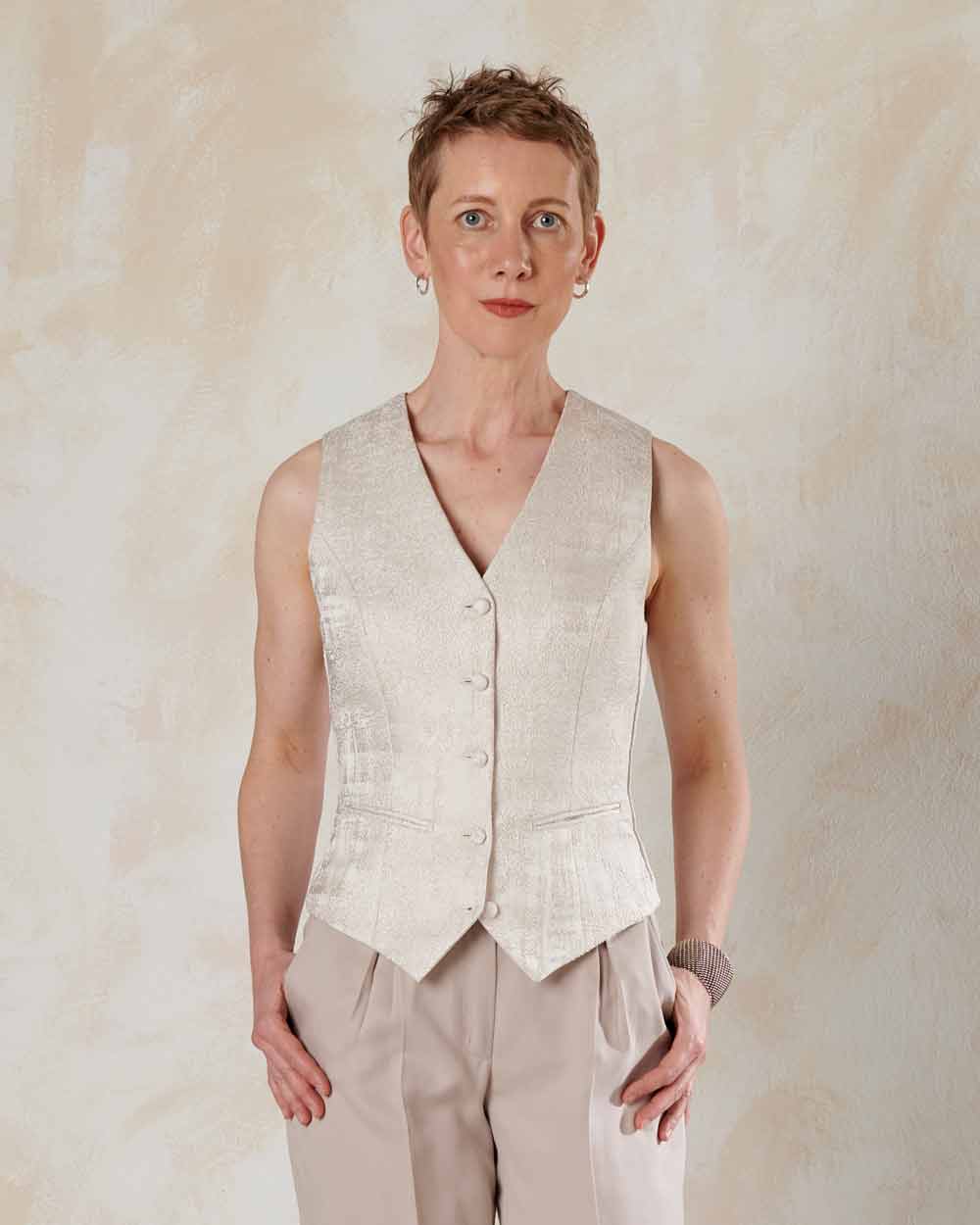 xuits-Frau mit kurzem Haar trägt beige Weste mit Knöpfen und helle Hose vor neutralem Hintergrund