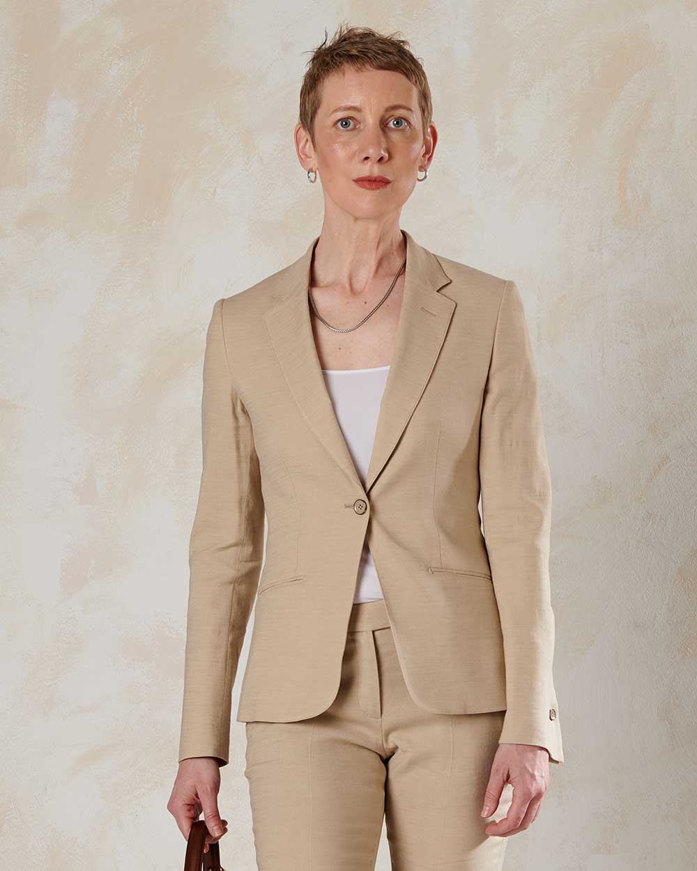xuits-Frau mit kurzem Haar in beigem Blazer, passender Hose, weißem Oberteil vor hellem Hintergrund.
