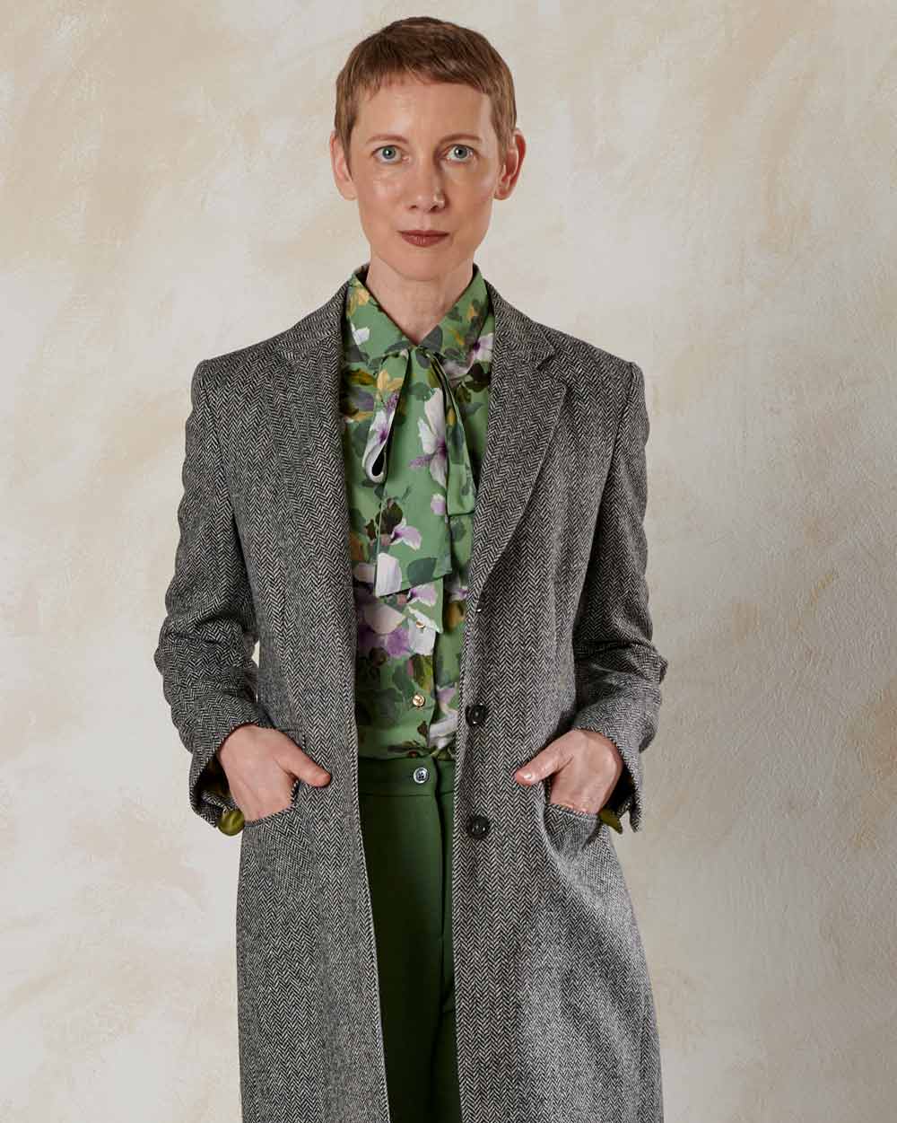 xuits-Frau mit kurzem Haar trägt grauen Mantel über grüner Bluse mit Blumenmuster vor heller Wand.