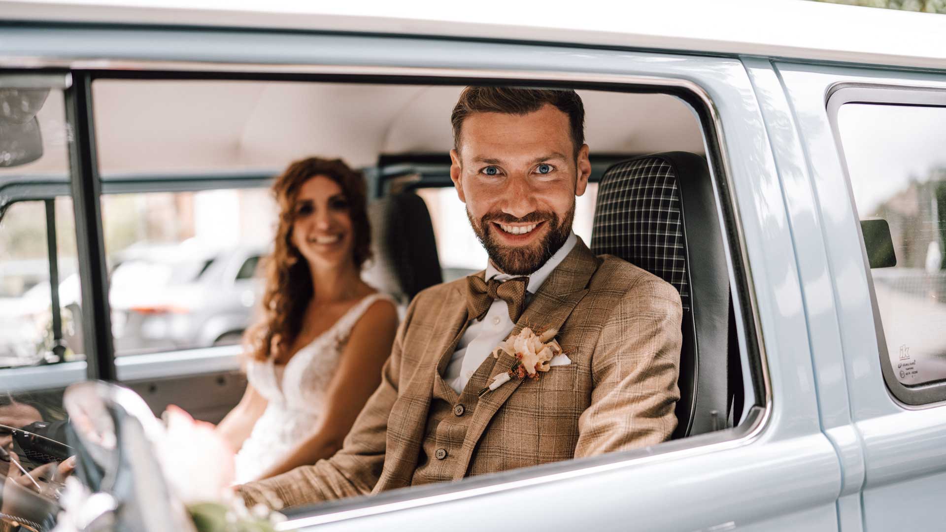 xuits -Brautpaar in Hochzeitskleidung sitzt lächelnd in einem Auto.