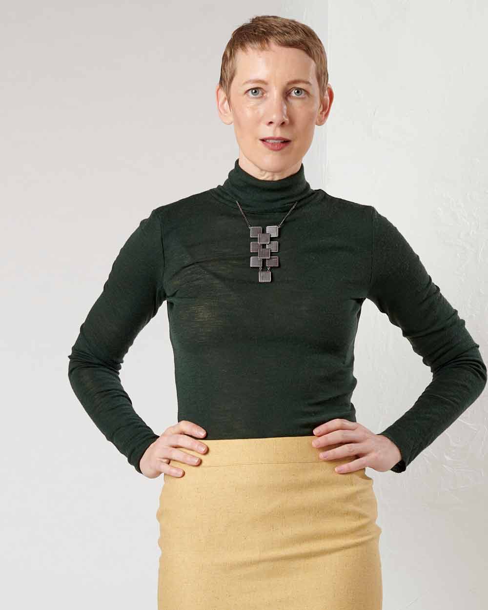 xuits -Frau mit kurzem Haar, schwarzem Rollkragenpullover, gelbem Rock und schwarzer Halskette vor hellem Hintergrund.