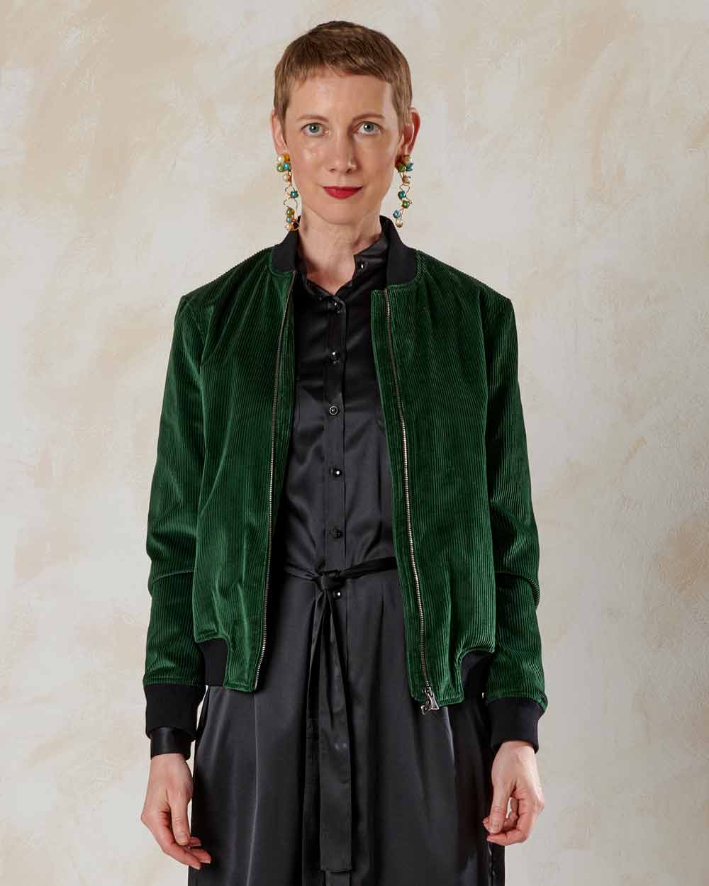 xuits-Frau mit kurzen roten Haaren in grüner Samtjacke und schwarzem Satin-Kleid vor hellem Hintergrund.